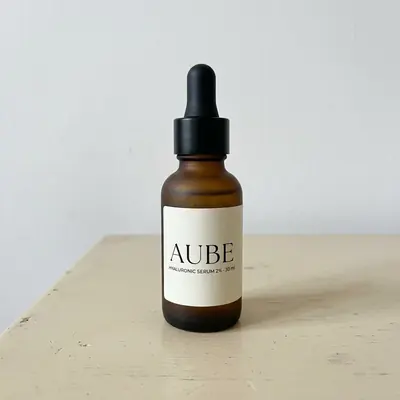 Photo fournisseur de départ : le sérum AUBE en flacon compte-gouttes ambré, prise au téléphone sur une table, sans retouche